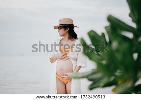 Beautiful asian young pregnant woman wearing orange bikini is standing in shallow water at Phangan island in evening time. Selective focus and split color.