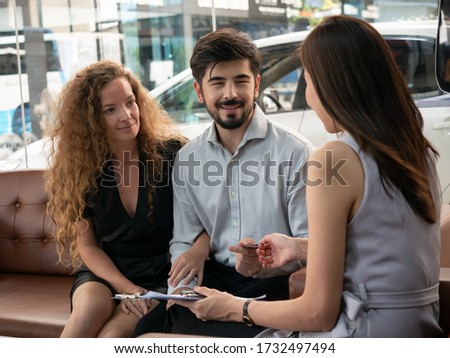 Confident young salesman explaining the car features to cacausian couple at car salon. Smiling couple buying a new car at car showroom.
