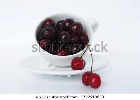 Photo of cherries in a white tea mug and saucer on a white background.
