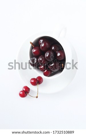 Photo of cherries in a white tea mug and saucer on a white background.

