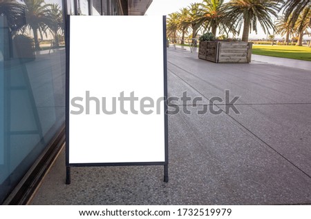 Blank white advertising stand/sandwich board mockup template. Background texture of clear street signage board placed outdoor on pedestrian sidewalk with some palm trees in a park as background.