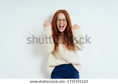 Exultant young redhead woman cheering and clenching her fists in excitement after receiving good news standing against a white wall with copy space Royalty-Free Stock Photo #1732591163