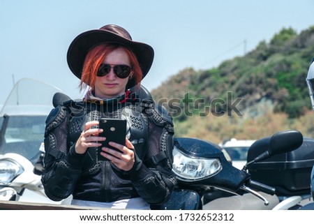 A girl in motorcycling outfit is standing by the road looking at the screen of a smartphone. Communication and navigation while traveling. Scooter in the background.