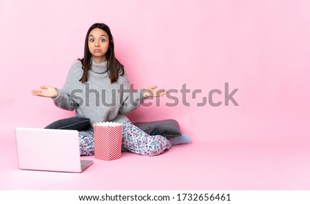 Young mixed race woman eating popcorn while watching a movie on the laptop making doubts gesture