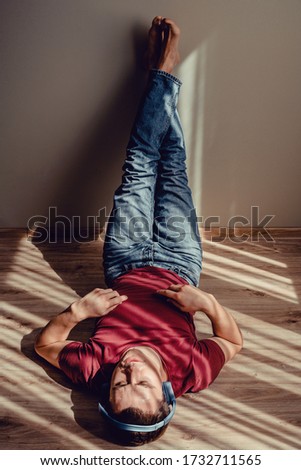 Carefree relaxed man in jeans, wine red t-shirt listening to music lying down on parquet floor, feet on wall. Natural light and shadow pattern stripes. Stress free concept. Blue wireless headphones