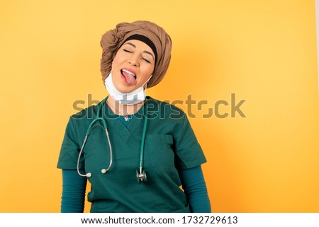 Beautiful muslim doctor woman with happy and funny face smiling and showing tongue. Wearing hijab, medical uniform and mask standing against gray studio background.