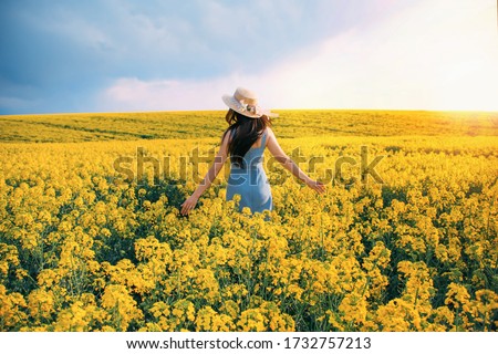 spring day. happy woman turned away back, walks run along yellow field rapeseed, hands raised to side enjoys nature blue sky. Girl brunette long black hair fly in wind. Jeans sundress straw hat flower Royalty-Free Stock Photo #1732757213