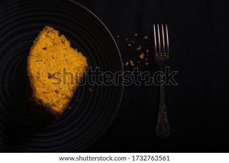 
A delicious slice of homemade carrot cake on a black plate accompanied by a fork. Black background. Aerial view.