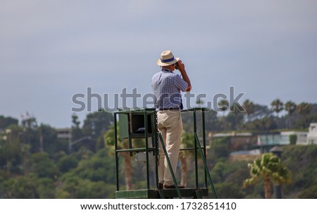 Picture of a man from behind on top of a lookout tower with looking through binoculars at the horizon.
