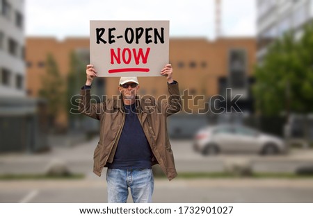 Protester with cap and sunglasses demonstrate against stay-at-home orders due to the COVID-19 pandemic carrying sign saying Re-open. Blurred buildings in the background.