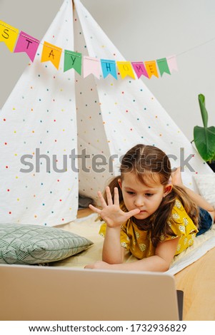 Tired little girl lying on a blanket, watching cartoons on a laptop without blinking. It's taking place in front of a hut, made with sticks and bedsheets. Stay at home flag garland hanging overhead.