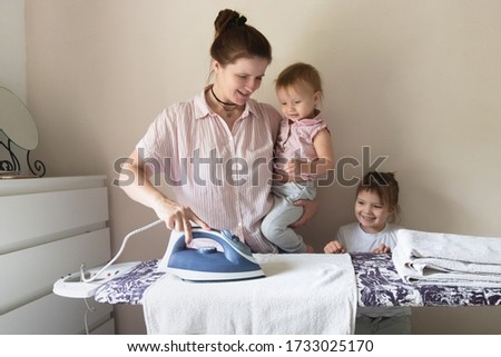 Caucasian mother with two emotional sibling children with an iron ironing linen on an ironing board, mother's assistants. Children help mom in household chores, ironing