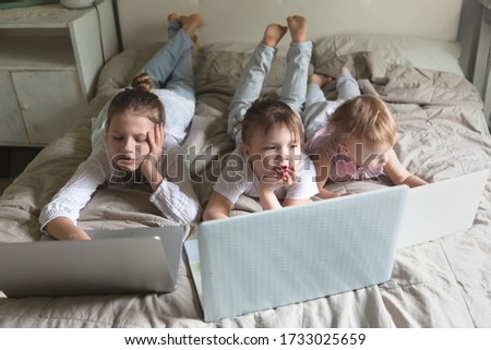 Three siblings children on a bed with laptops, concept digital addiction, home, distance learning online, tired children behind monitors