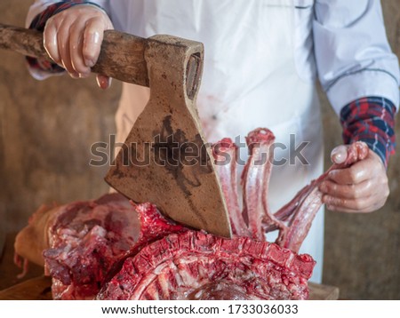 elderly man with a gray beard in cook suit and white apron cuts into pieces large piece of pork with knife and ax