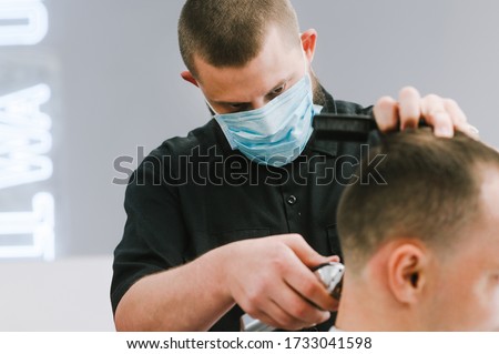 Professional barber in a medical gauze mask on the face cuts the client's hair with a clipper with a focused gaze. Hairdresser works in quarantine. Close up Royalty-Free Stock Photo #1733041598