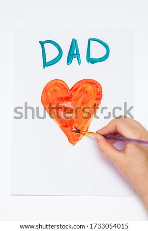 Top view of hand of child drawing red heart with word Dad greeting card on white paper. Family and Father's Day concept.