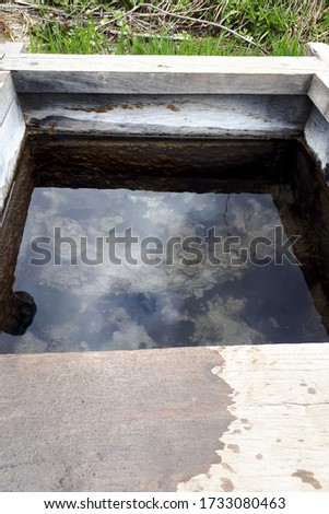 Wooden well with clear water in the countryside. Reflection of the sky with clouds in calm water. Vertical photo.