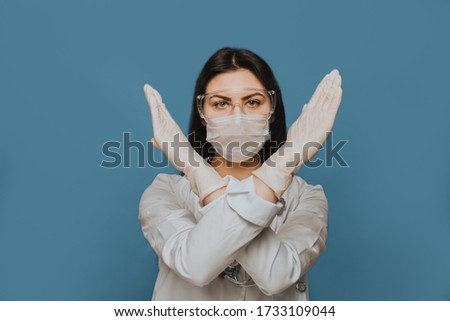 Nurse wearing protective glasses, a surgical mask and a medical gown crossed her hands shows gesture stop with meaning that we need to stop coronavirus pandemic. Covid-19 virus concept.