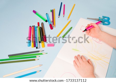 a small child draws with a pink felt-tip pen in an album, copy space, top view, blue background, pencils, wax crayons, scissors and the child's hands on the table