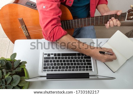 Aerial shot of a man whose face cannot be seen at home taking guitar lessons online and pointing in a notebook. Activities during quarantine