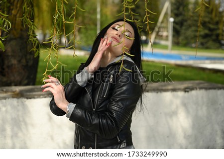 Model posing against the backdrop of a city park. Photo in spring weather outdoors caucasian fashionable brunette girl in a black jacket.