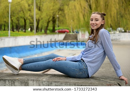 Photo of a young pretty girl with a smile in a blue shirt and jeans. Model posing sitting in spring park outdoors in the city.