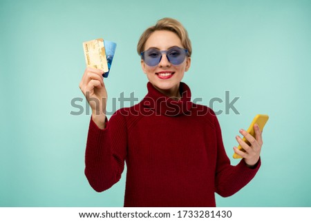 Photo of pleased young woman posing isolated over blue wall background using mobile phone holding debit card. Focus on cards