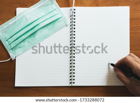 picture of hand using pen writing phase on the diary book with surgical face mask on top on the sooden table. covid-19, coronavirus and new normal lifestyle concept