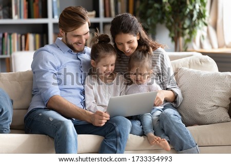 Happy young Caucasian family with small daughters relax on couch at home watching funny video on laptop, smiling parents with little girls children enjoy home weekend using computer gadget together