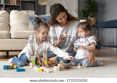 Loving young Caucasian mother sit on floor in living room playing building bricks with little daughters, caring mom or nanny construct with wooden block with small girls children at home together Royalty-Free Stock Photo #1733355050
