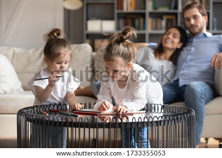 Little preschooler girls drawing painting with colorful pencils at home, young Caucasian parents relax on sofa in background, happy family rest in living room at home with small daughters on weekend