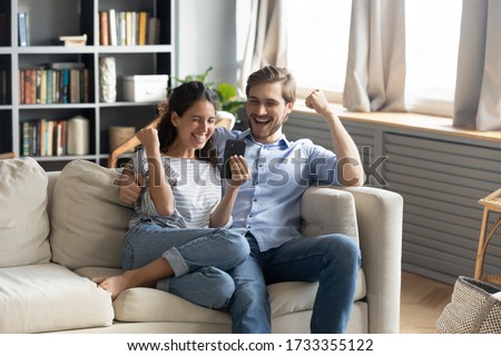 Overjoyed young Caucasian couple sit relax on couch in living room triumph winning online lottery on cellphone, happy man and woman feel excited reading good unexpected pleasant news on smartphone Royalty-Free Stock Photo #1733355122