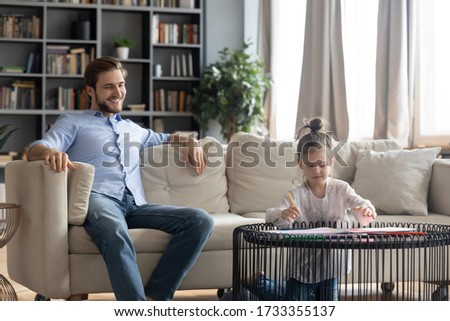 Smiling young Caucasian father sit relax on couch in living room watch little daughter drawing in album with colorful pencils, happy dad enjoy family weekend with small girl child at home together