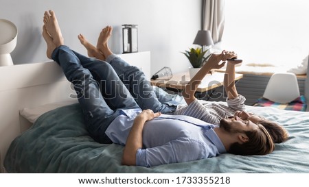 Happy millennial Caucasian couple lying relaxing in comfortable bed at home using smartphone together, loving young man and woman rest in bedroom make self-portrait picture on modern cellphone gadget
