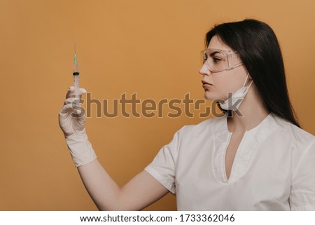 Nurse in protective glasses and a dressing scrubs, holding a syringe is about to inject a patient with coronavirus, over yellow background. Covid-19 concept.