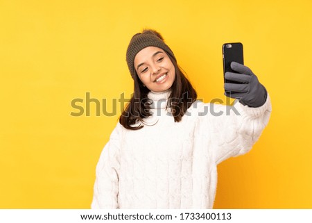 Young woman with winter hat over isolated yellow background making a selfie