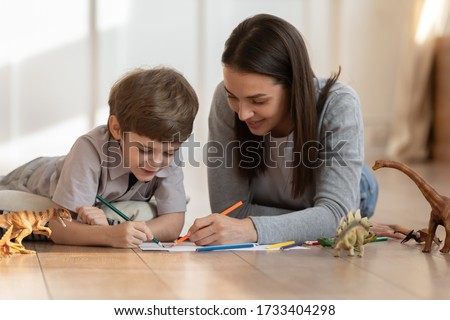 Loving young mom or nanny lying on warm floor drawing in album with little boy child, smiling mother have fun play paint on paper with small son on home family weekend, children creativity concept