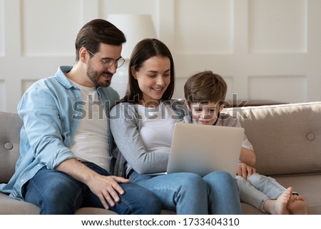 Happy young family with small preschooler son sit relax on sofa in living room watch video laptop gadget together, smiling parents with little boy child using computer at home, enjoy weekend