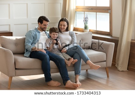 Happy young Caucasian family with son sit relax on sofa in living room using modern smartphone gadget together, smiling parents rest on couch at home with small boy child, browsing cellphone device