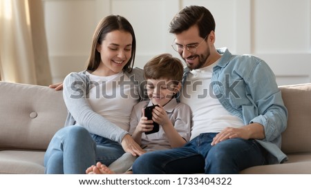 Smiling young parents with small preschooler son sit relax on couch in living room watch funny video on cell, happy Caucasian family with little boy child rest on sofa ta home using smartphone gadget