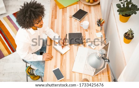 Copywriter working from Home. Creative young man writes notes in note pad looking at laptop. Arabic guy student in eyeglasses working or study. High angle view.