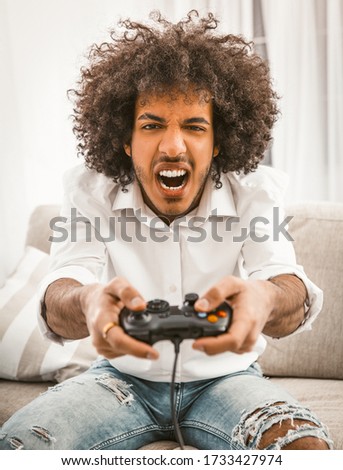 Screaming gamer shoots or attacks in computer game. The concept of emotions. Young Arab playing computer game sitting at sofa in home interior.