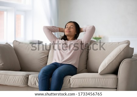 African ethnicity millennial woman in casual clothes puts hands behind head closed eyes resting lean on sofa. Daydream, enjoy fresh conditioned air at modern flat, no stress, reducing fatigue concept Royalty-Free Stock Photo #1733456765