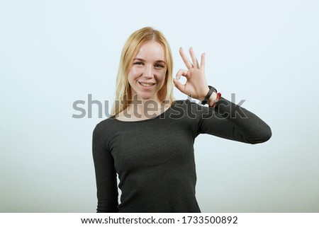Young Blonde Woman In Black Sweater With Stylish Watch On White Background, Happy Girl Smiles, Makes Ok Sign With Her Fingers. The Concept Of Good, Successful People