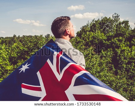 Attractive man holding Australian Flag on blue sky background on a clear, sunny day. View from the back, close-up. National holiday concept
