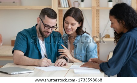 Smiling young family clients make purchase deal sign insurance contract at meeting with african american estate agent. Happy millennial couple reading document about buying house on loan.