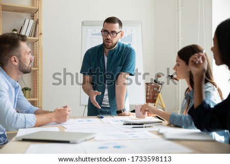 Confident bearded businessman with glasses presentation new project in coworking boardroom at company meeting. Serious man coach auditor speaks about business on flip chart graphs background.
