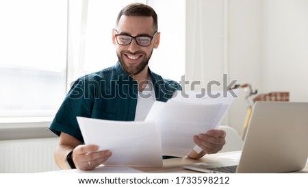 Happy handsome businessman holding and reading documents with good news near laptop. Young smiling bearded man, student rejoice and celebrate successful distance deal for work. Royalty-Free Stock Photo #1733598212