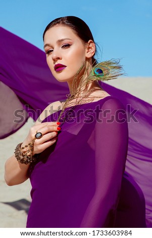 Amazing beautiful brunette woman with the Peacock feather in purple fabric in the desert. Oriental, indian, fashion, style concept