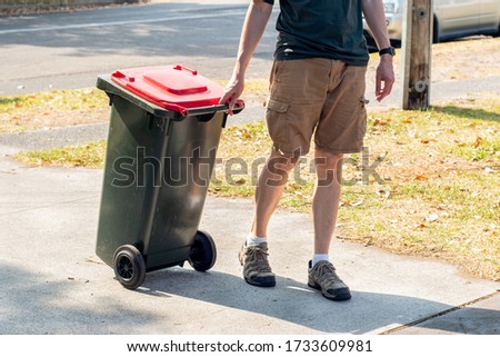 A man dragging the household wheelie red bin with general waste on the street for council gargbage collection. Waste management concept. Royalty-Free Stock Photo #1733609981